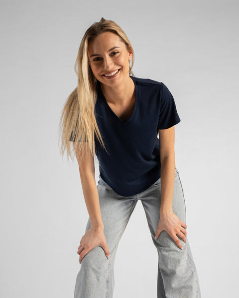 A studio portrait of a blonde woman leaning forward with a big smile, looking directly at the camera. Her hands are resting on her thighs just above her knees. She is wearing a regular fit, dark navy blue short-sleeved v-neck t-shirt and light-wash blue jeans against a plain light grey background.