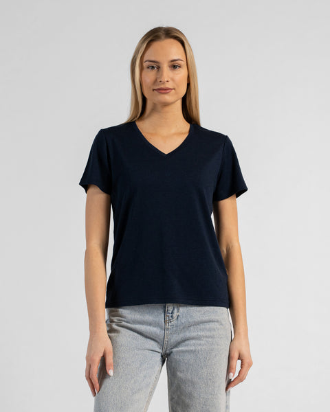 A studio portrait of a blonde woman standing and looking directly at the camera with her arms relaxed by her sides. She is wearing a regular fit, dark navy blue short-sleeved v-neck t-shirt and light-wash blue jeans. The background is a solid light grey.