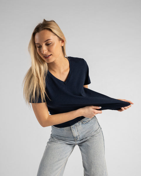A studio portrait of a blonde woman playfully stretching the hem of her dark navy blue short-sleeved V-neck t-shirt outward with both hands. She is wearing light wash denim jeans and looking down and to the left against a plain light grey background.