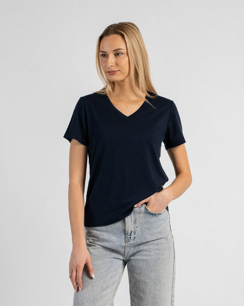A studio portrait of a blonde woman standing and looking slightly off to the left against a plain light grey background. She is wearing a dark navy blue short-sleeved V-neck t-shirt and light wash denim jeans, with one hand resting in her front pocket.