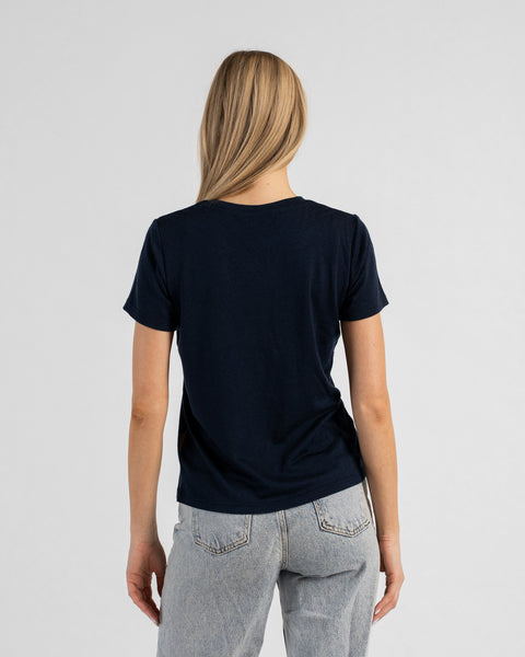 A back view studio portrait of a blonde woman standing against a plain light grey background. She is wearing a dark navy blue short-sleeved V-neck t-shirt and light wash denim jeans, with her straight blonde hair falling down her back.