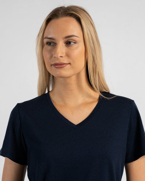 A studio portrait of a blonde woman standing and looking off to her right against a plain light grey background. She is wearing a dark navy blue short-sleeved V-neck t-shirt.