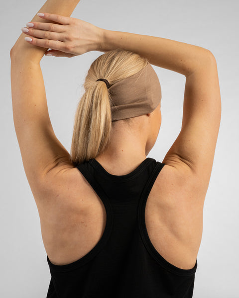 A woman viewed from behind, wearing a black racerback tank top and a beige headband. Her blonde hair is pulled into a ponytail, and she has her arms raised, hands resting behind her head.