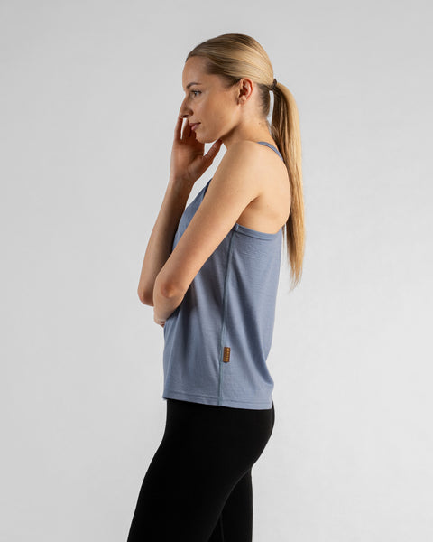 A profile shot of a woman standing against a white background, wearing a dusty blue sleeveless top with a small brown logo tag on the side and black leggings. Her blonde hair is pulled back in a ponytail, and she is gently touching her face with one hand.