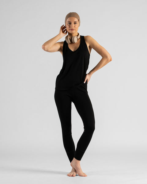 A woman kneeling barefoot on a white background, wearing a black sleeveless top, black leggings, and a beige headband. Beige headphones rest around her neck as she poses with one hand on her hip, looking at the camera.
