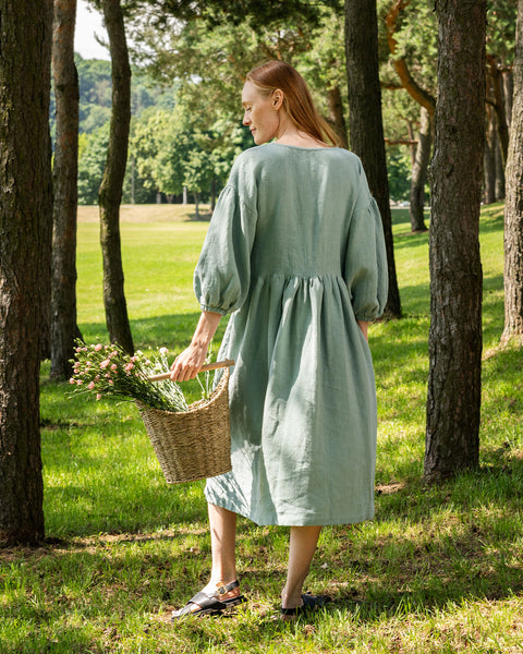 A person with light skin and light brown hair stands facing away from the camera in a grassy, wooded area, holding a woven basket with flowers. They are wearing a long, mint green linen dress with puffed sleeves and sandals.