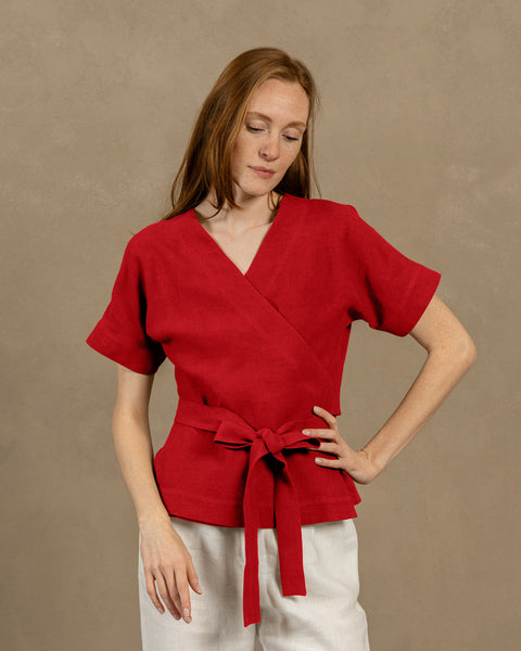 A fair-skinned person with long, auburn hair, wearing V-neck linen wrap top with short, wide sleeves and light-colored pants. They are holding the ends of the top's tie in front of them, forming a bow.