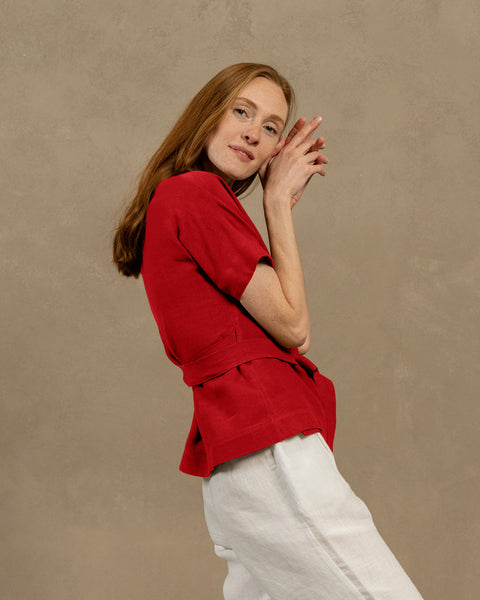 A fair-skinned person with long, auburn hair, wearing pure red V-neck linen wrap top with short, wide sleeves and light-colored pants. They are holding the ends of the top's tie in front of them, forming a bow.