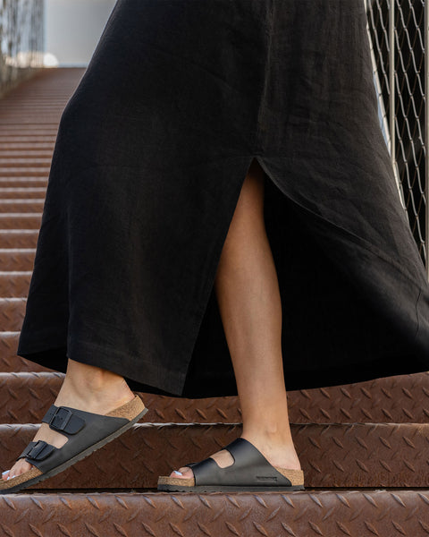 A close-up shows the lower portion of a person wearing a long pure black menique dress with a side slit, revealing their legs and feet in black two-strap sandals with cork soles. They are standing on textured metal stairs with a wire mesh railing visible in the background. The sandals appear to be Birkenstock Arizona style.