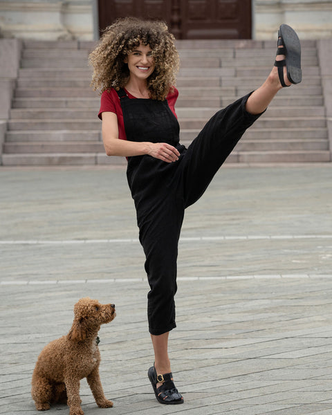 Beautiful woman with curly hair posing in the city wearing pure black linen jumpsuit Nicci, royal cherry merino wool top. Posing with a brown poodle dog.