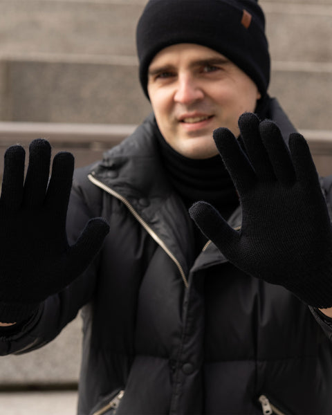 A smiling man in a black winter beanie and a black puffer jacket extends both hands towards the viewer, palms open, showcasing his black knitted gloves. He is looking directly at the camera. The background is a blurred view of outdoor stone steps.