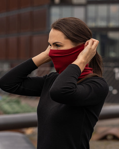 A woman outside in a city, wearing the menique unisex neck gaiter in royal cherry. She is pulling it up over her nose and even ears. She also wears a black merino long sleeve top.