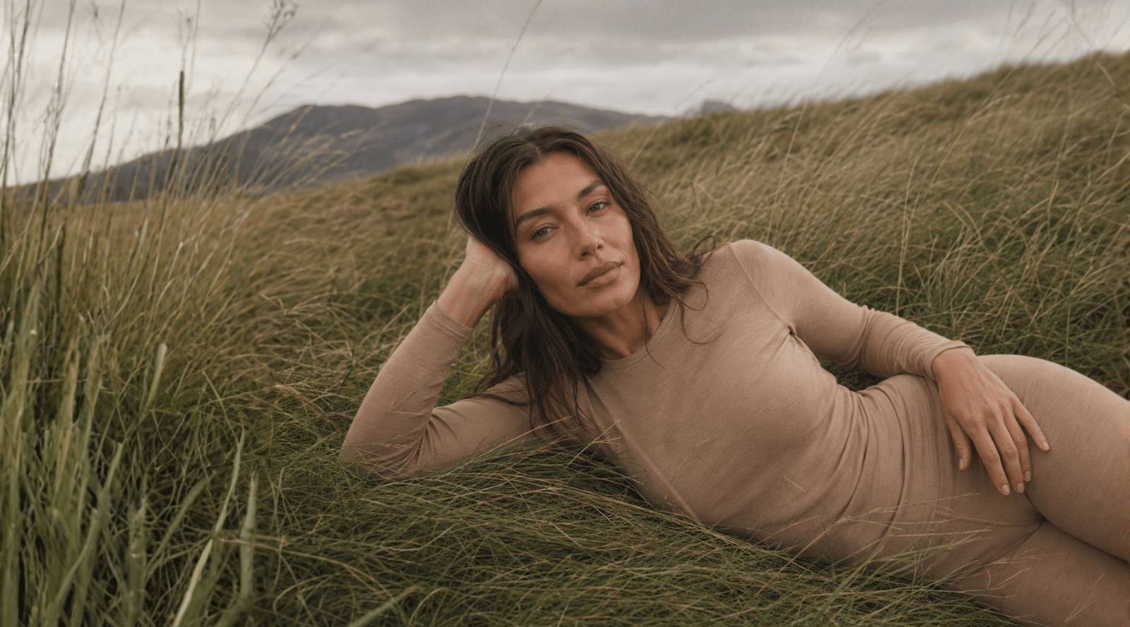 A woman wearing a beige long-sleeved top resting her head on her hand while lying in tall, green grass.