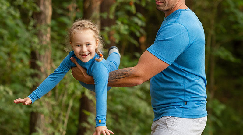 In the photo you can see a man holding his baby girl, who is wearing a matching bright blue set made from organic and lightweight 100% Merino wool.