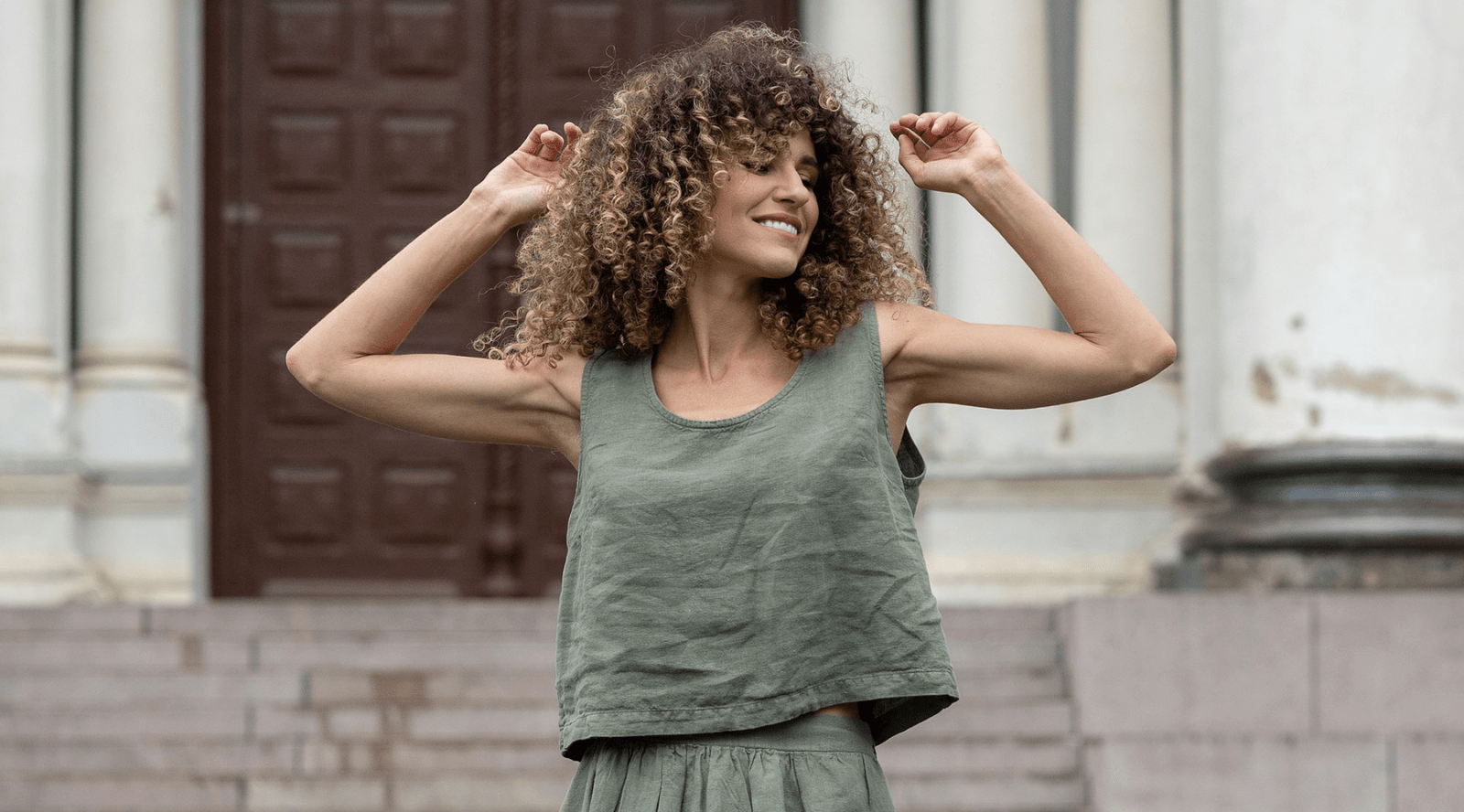 a woman standing by a building, wearing a full black linen set, that includes black pants and a black linen button-up shirt. She is smiling and has curly hair.