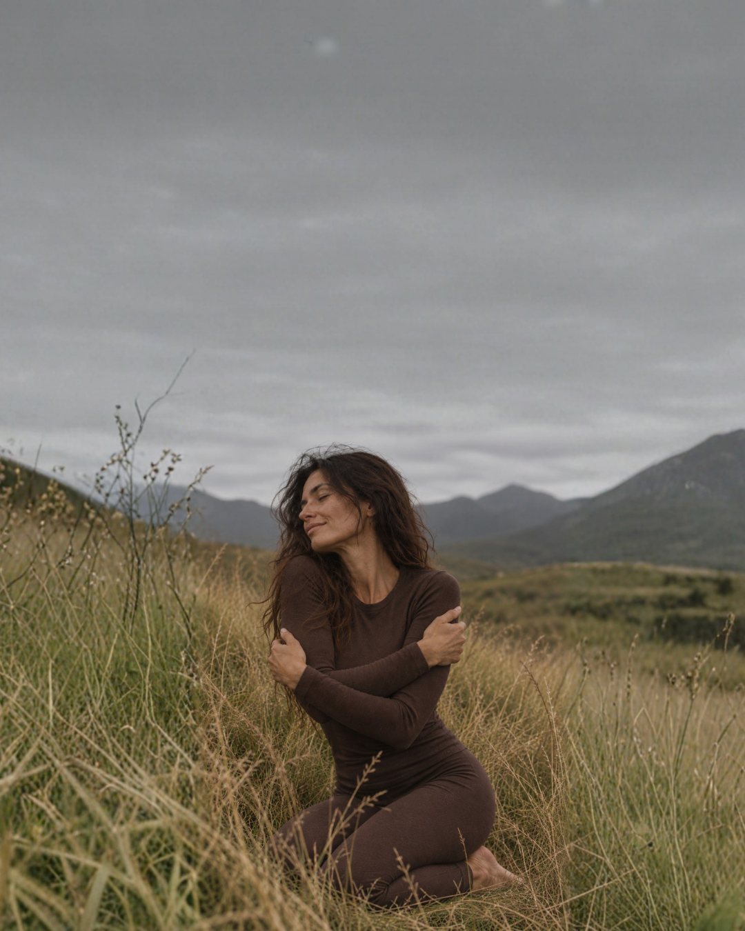 A woman kneels peacefully in a field of tall grass with her eyes closed and her arms crossed over her chest. She is wearing a brown long-sleeved thermal set, framed by distant mountains and a cloudy gray sky.