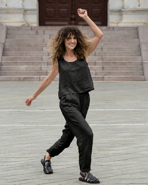 A joyful woman with curly hair mid-stride in a stone plaza, with one arm raised playfully in the air. She is wearing a black sleeveless linen top, matching cropped pants, and black sandals.
