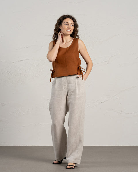 A smiling woman with long, curly brown hair wears a sleeveless linen crop top with side ties and full-length, wide-leg natural linen pants. She stands with one hand in her pocket, looking slightly to the side, against a textured light gray wall and a dark gray floor.