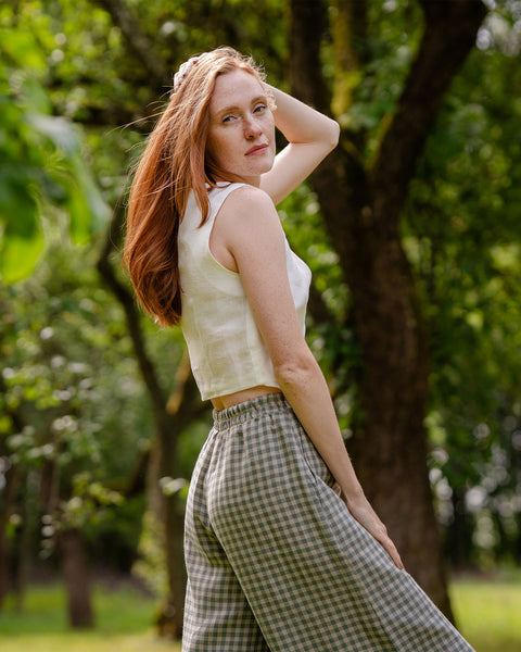 The model in the photograph is wearing mid-rise, light green and white gingham pattern wide leg linen pants. She has matched them with a white sleeveless linen crop top. Posing sideways, she reveals the elastic waist at the back of the pants.