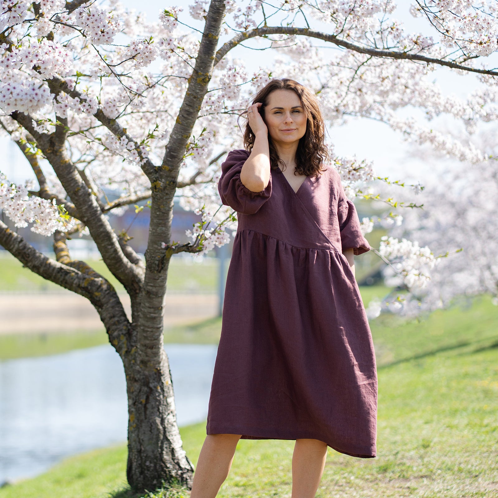 A smiling woman with dark, wavy hair stands outdoors next to a blooming cherry blossom tree. She is wearing a knee-length, shadow purple linen wrap dress with elbow-length sleeves. Her right hand is raised to her face. In the background, there is a body of water and green grass under a bright sky.
