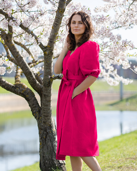A light-skinned person with shoulder-length brown hair is standing near a blossoming tree, looking towards the left. They are wearing a hot pink wrap dress with short puff sleeves and a tied waist, with one hand in a pocket and the other gently touching their hair. The background shows a blurred landscape with hints of green grass and water.