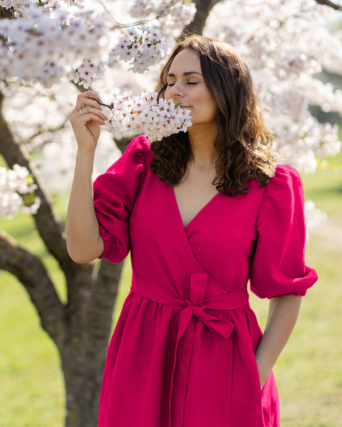 A light-skinned person with shoulder-length brown hair has their eyes closed and appears to be smelling a cluster of white blossoms held in their right hand. They are wearing a hot pink wrap dress with short puff sleeves and a tied waist, and their left hand is in a pocket. A blossoming tree and a blurred green and tan background are visible.