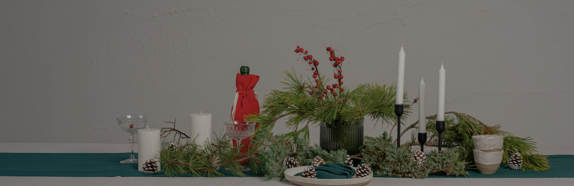 A Christmas dining table decorated with candles, glasses, a wine bottle inside a festive linen bag and natural elements, such as pinecones and tree branches. The table is covered with a pure white tablecloth and a dark green table runner.