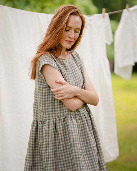 The model is wearing a cap sleeve, knee-length gingham dress in a light green and white checkered pattern, standing with her arms crossed outdoors near a clothesline with white garments hanging in a garden.