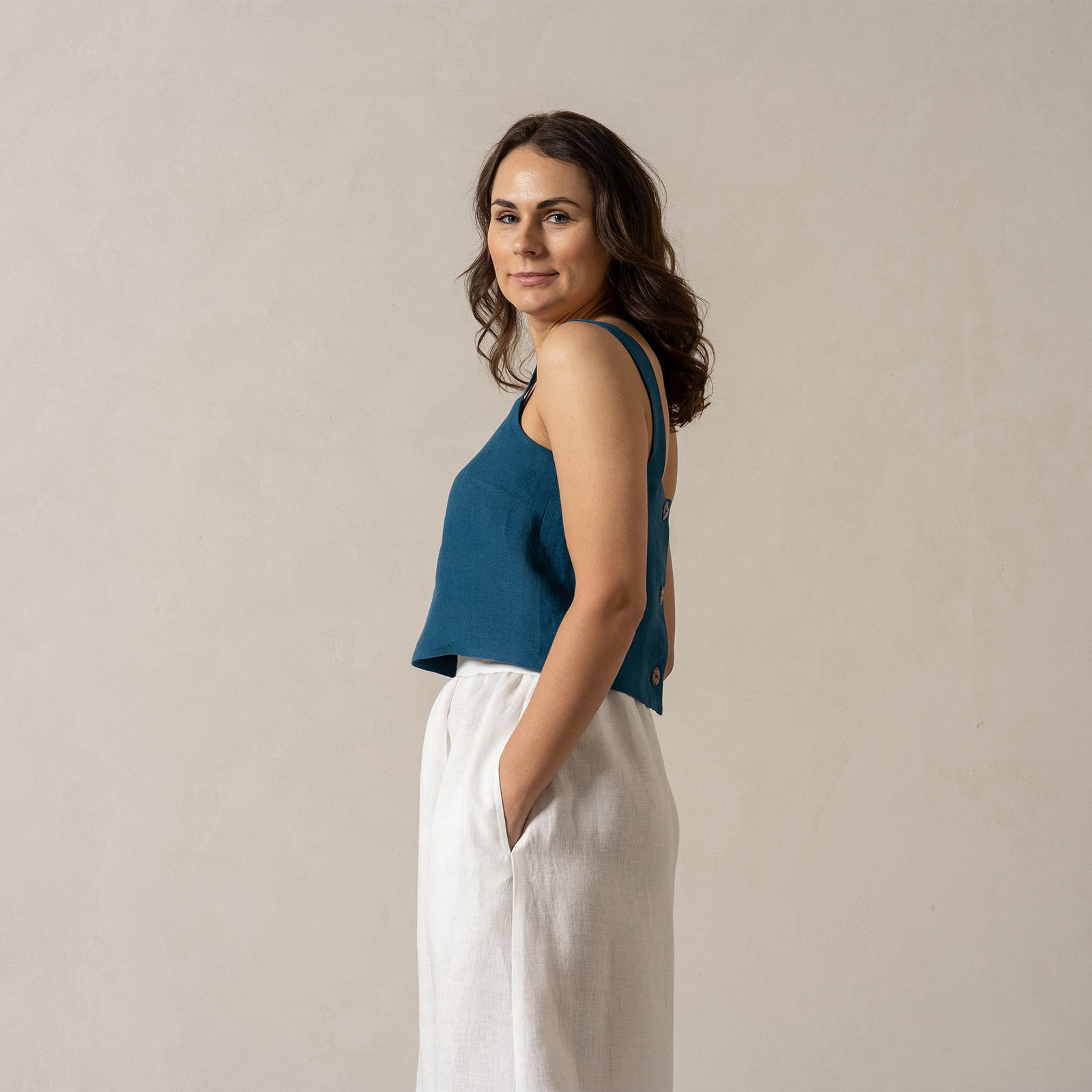 A woman with medium-toned skin and dark hair looks directly at the camera with a neutral expression. She is wearing a teal, sleeveless square-neck linen top and a pure white, menique
 linen skirt, with her hands in the skirt's pockets. The background is a plain, light beige color.