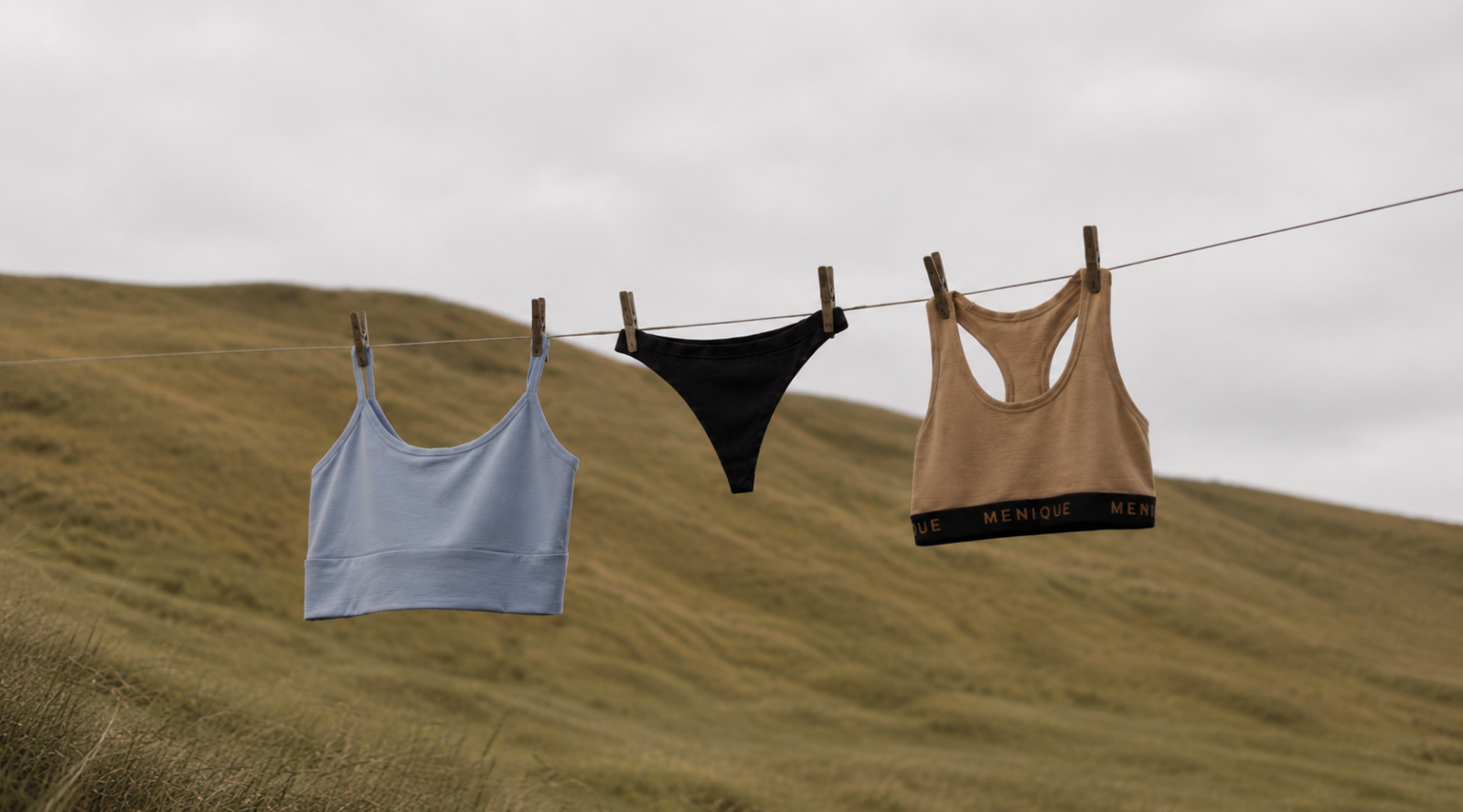 Three pieces of women's underwear or activewear hanging on a clothesline outdoors against rolling grassy hills under a cloudy sky: a light blue crop top, a black bikini bottom, and a beige racerback crop top with black trim.