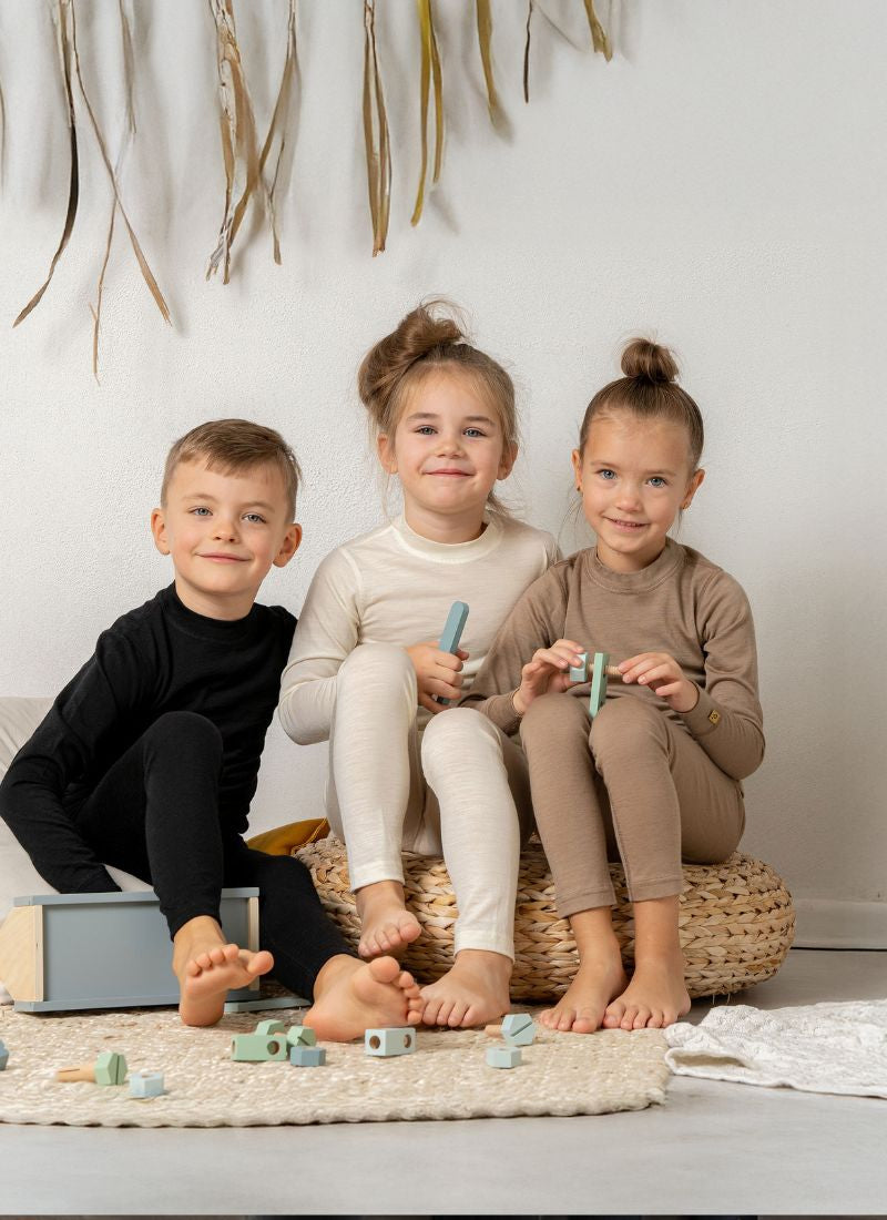 Three children sitting on a woven stool with toys on a light-colored floor.