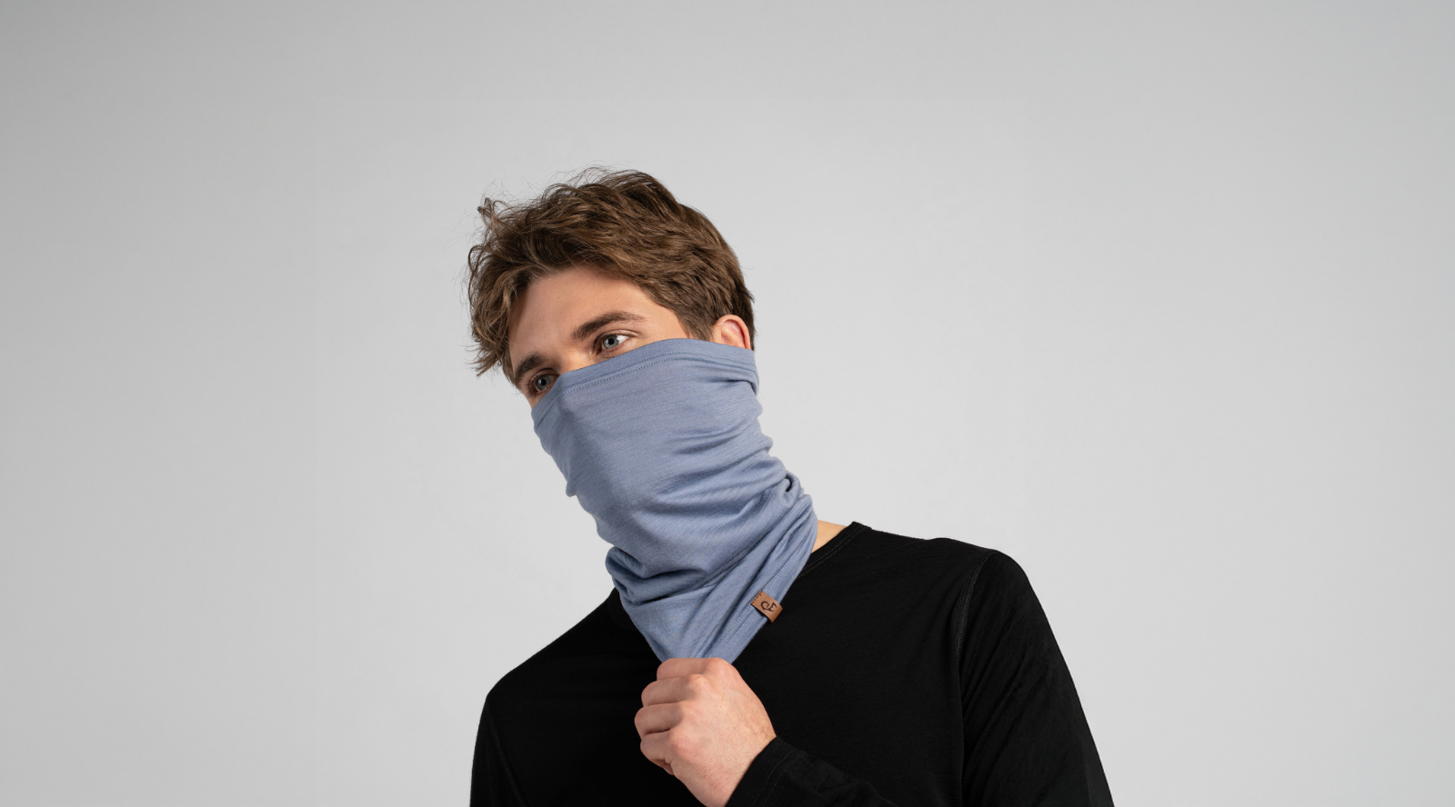 A man with wavy brown hair models a light blue knit neck gaiter pulled up to cover his nose and mouth. He wears a black long-sleeved shirt against a seamless light grey background, lightly holding the bottom edge of the gaiter.