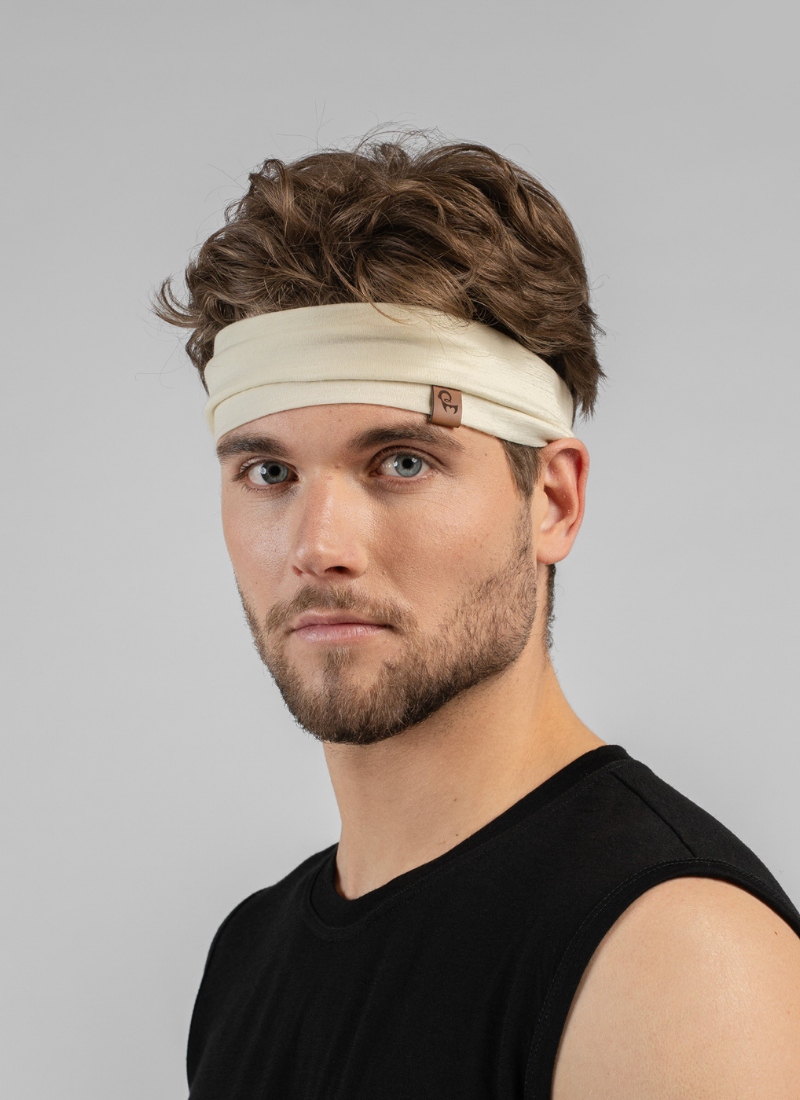 A man with wavy brown hair looks at the camera while wearing a wide, cream-colored knit headband. He is dressed in a simple black sleeveless top against a light grey studio background.