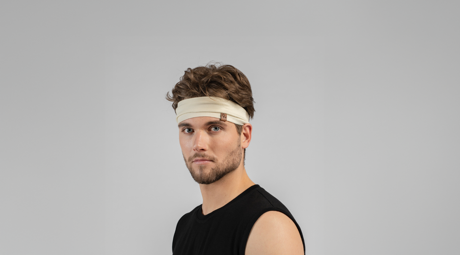 A man with wavy brown hair looks at the camera while wearing a wide, cream-colored knit headband. He is dressed in a simple black sleeveless top against a light grey studio background.