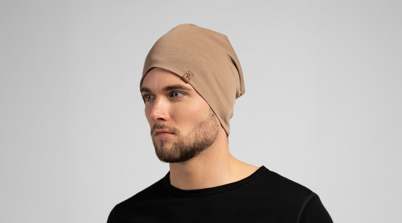 A bearded man looks slightly off-camera while wearing a slouchy, light brown knit beanie. He is dressed in a black crew-neck top against a seamless light grey studio background.