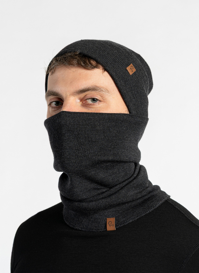 A man stands against a light grey studio backdrop, wearing a dark grey knit beanie and a matching neck gaiter pulled up over his lower face. He is dressed in a simple black long-sleeved top.