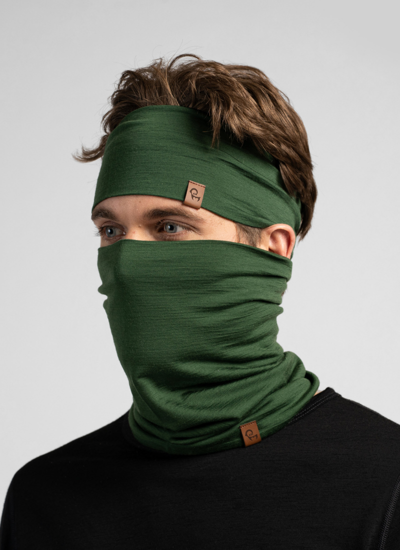 A man with brown hair models a matching dark green knit headband and neck gaiter pulled up over his nose and mouth. He wears a black top against a seamless light grey studio background.