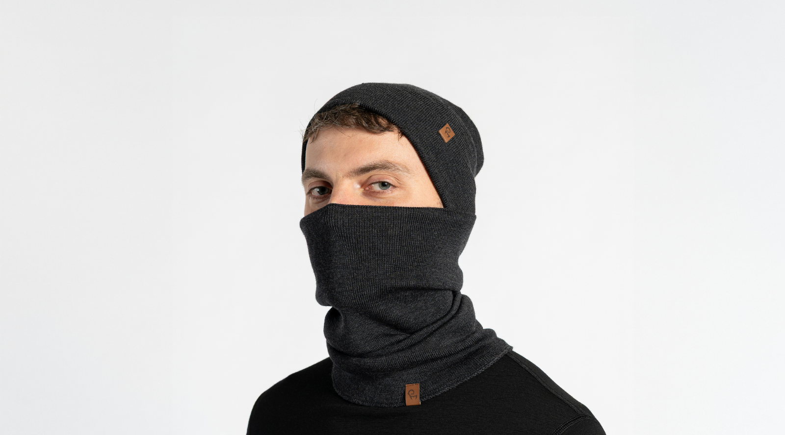 A man stands against a light grey studio backdrop, wearing a dark grey knit beanie and a matching neck gaiter pulled up over his lower face. He is dressed in a simple black long-sleeved top.