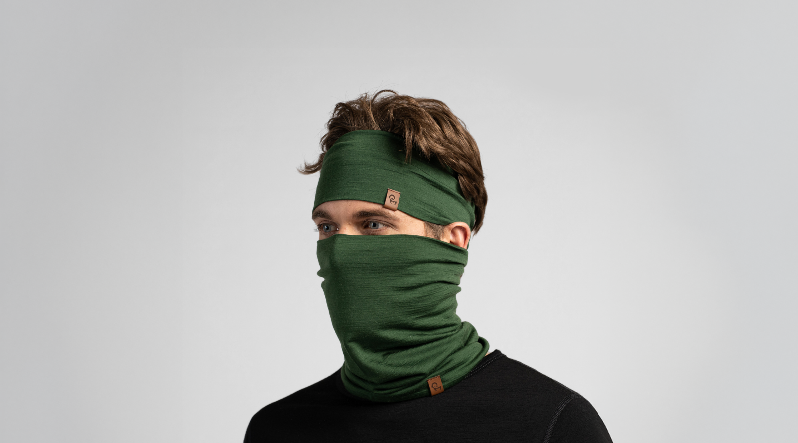 A man with brown hair models a matching dark green knit headband and neck gaiter pulled up over his nose and mouth. He wears a black top against a seamless light grey studio background.