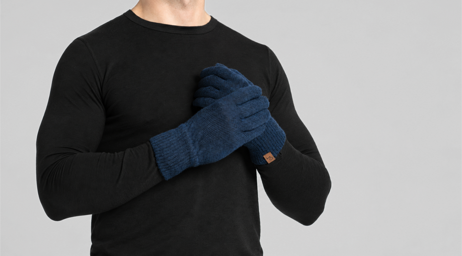 A person wearing a black long-sleeved shirt holds their hands crossed over their chest. They are wearing dark navy blue knitted gloves featuring a small brown logo tag on the cuff.