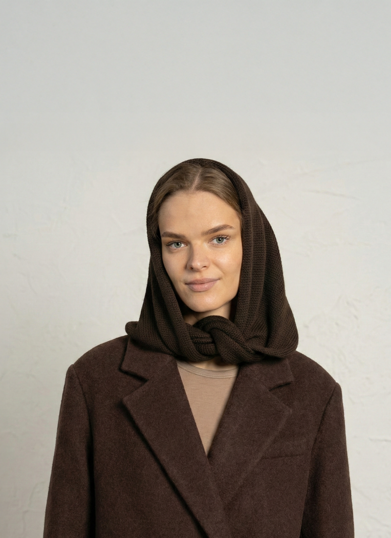 A blonde woman wearing a heavy dark brown coat over a tan top smiles softly in front of a textured off-white wall. A dark brown knit hood covers her head and wraps comfortably around her neck.