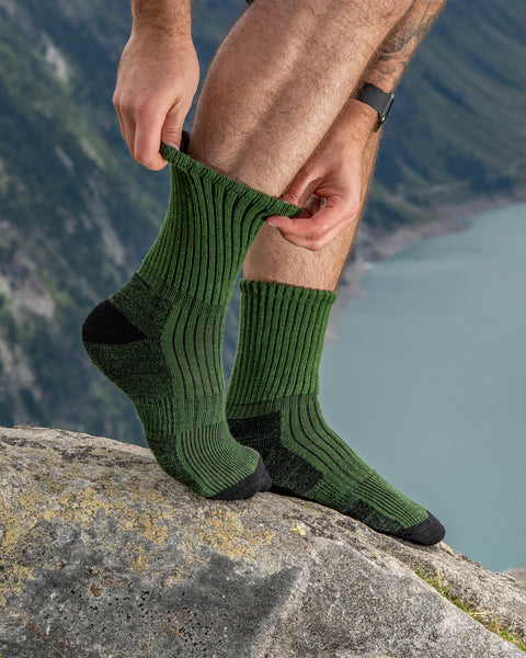 Hiker adjusting green ribbed Merino hiking socks, sitting on a rock above a turquoise mountain lake.