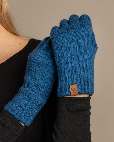 A close-up shot of a person wearing menique brand vibrant blue, ribbed knit gloves. The person is wearing a dark long-sleeved top, with the right hand holding the left wrist, showcasing the gloves&#39; fit and the small, rectangular tan leather menique tag on the cuff of the left glove. The background is a solid, neutral gray.