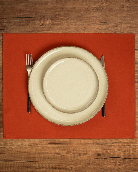 The menique linen placemat in cinnamon red color. There are two porcelain plates in different sizes on top of it, and cutlery by the sides of them. There is a wooden tabletop visible in the background.