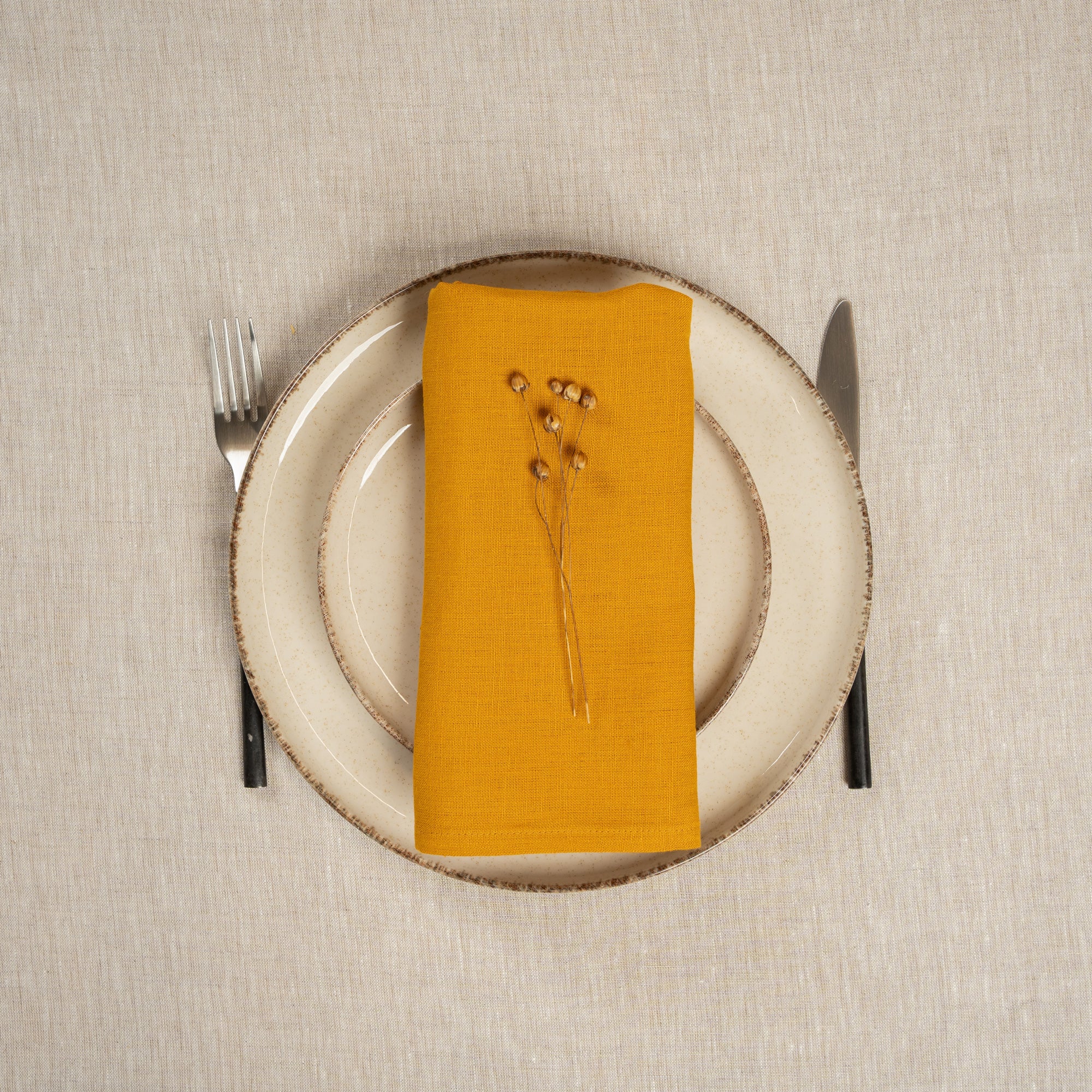 The menique linen napkin in spicy yellow color, put on top of two stacked plates . There are cutlery items by the sides of the plates. The background is a natural color linen tablecloth.