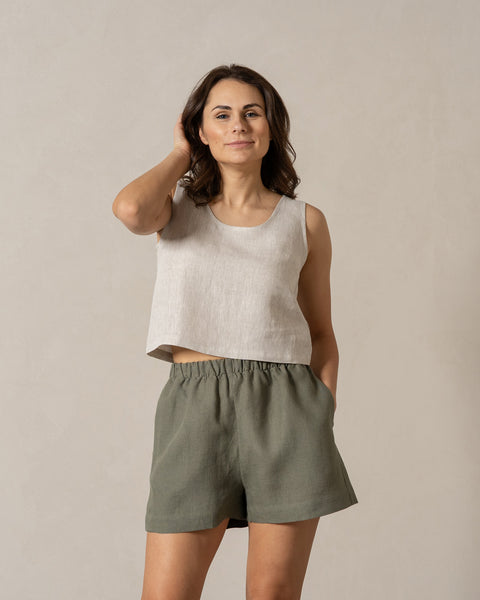 A woman with dark hair wearing 100% linen shorts in the color stone green and linen cross back crop top in the color natural. Her one hand is in the pocket while the other is touching her hair. The background of the photo is in natural, nude colors.