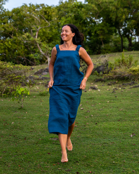 A woman with curly black hair runs barefoot uphill on a lush, green hill. She wears a cobalt blue, sleeveless menique dress that flows gently behind her.