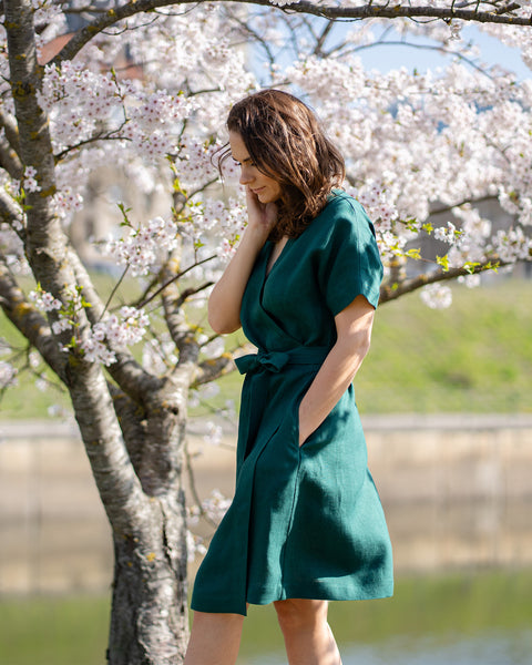 A woman with brown hair, wearing a dark green, short-sleeved wrap dress, stands outdoors under a tree with white blossoms.