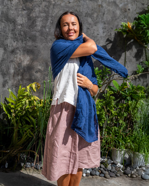 A person standing outdoors wearing a storm blue menique linen scarf draped over the shoulders, with a beige linen dress. The image captures a side profile of the person against a backdrop featuring a gray wall and greenery.