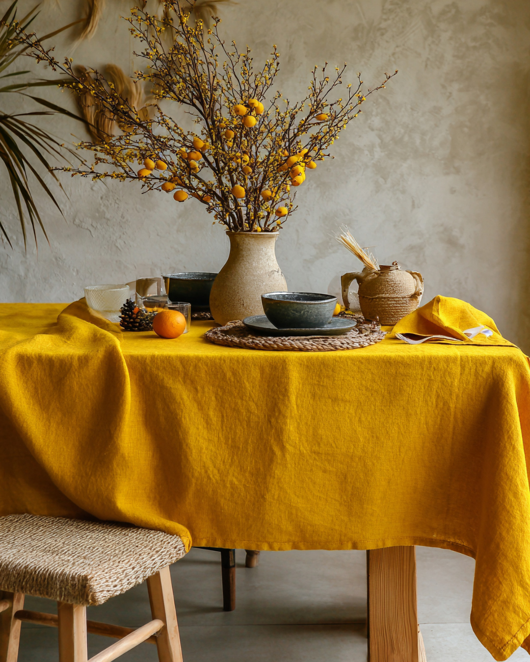 Dining table covered with a mustard yellow linen tablecloth, decorated with ceramic tableware, dried branches with yellow accents, and natural textures, showcasing warm, earthy home textile styling.